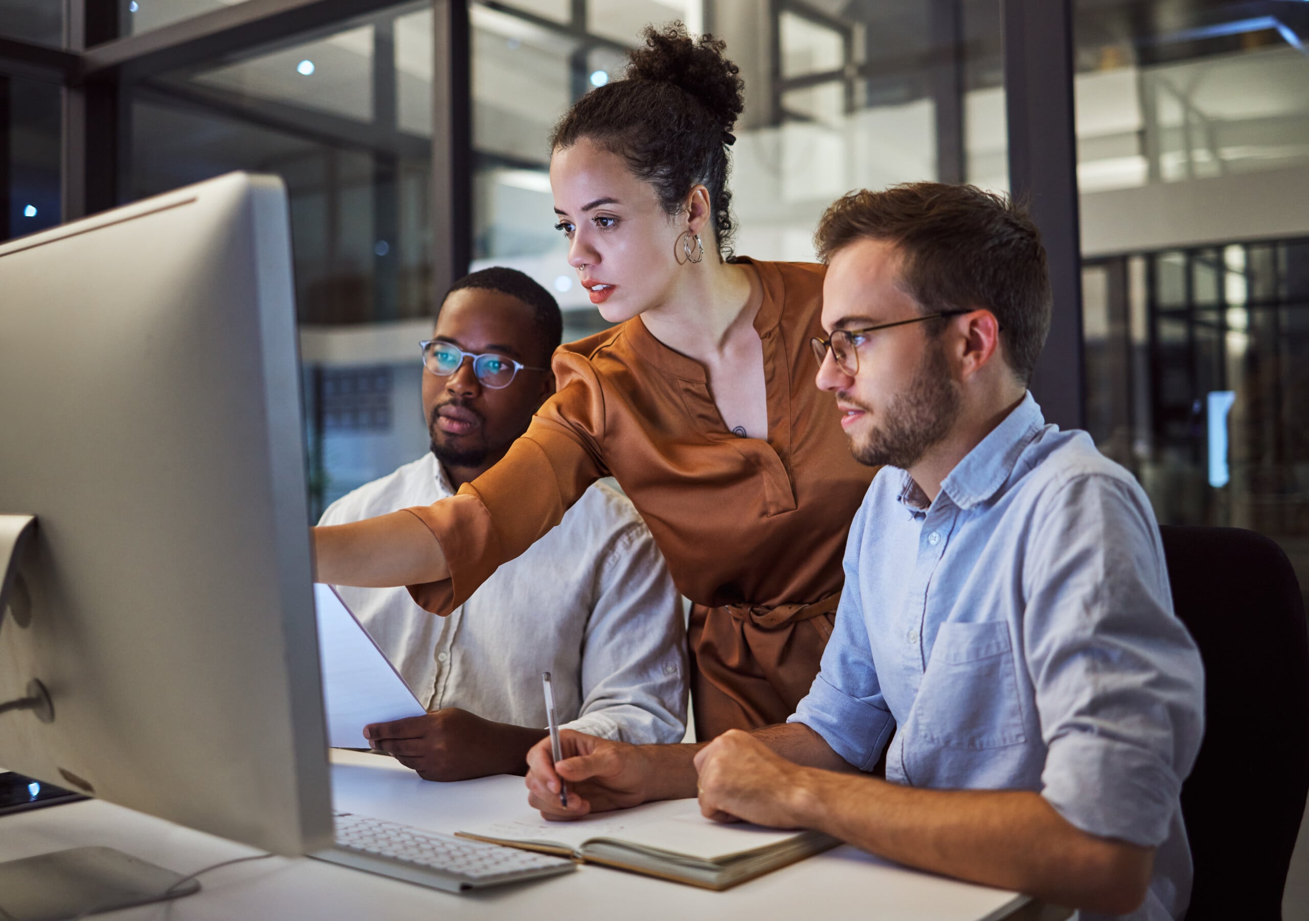 Two men sat at desk looking at screen, woman overlooking