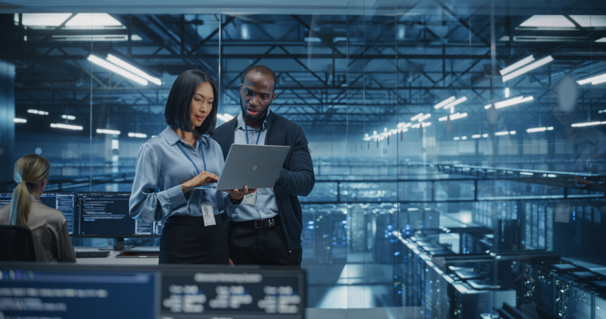 Male DevOps Engineer and Female IT Consultant Work on a Laptop Computer Standing in an Office Above the Supercomputer