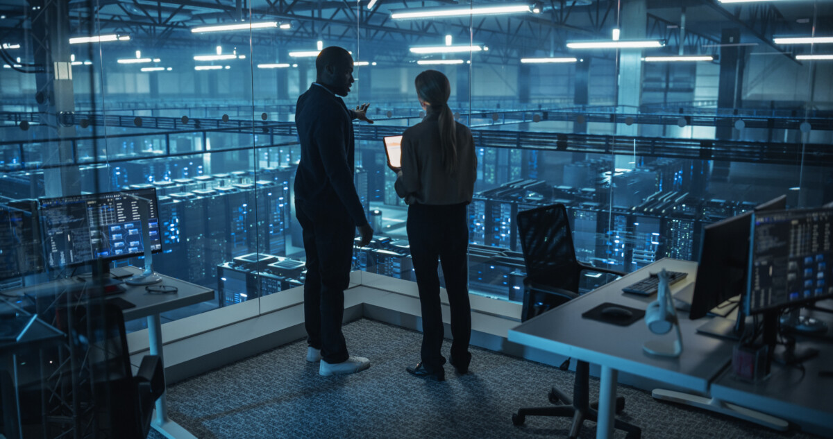 Male DevOps Engineer and Female IT Consultant Work on a Tablet Computer Standing in Office Above the Rows of Servers in a Data Center Working on Research and Development of Artificial Intelligence