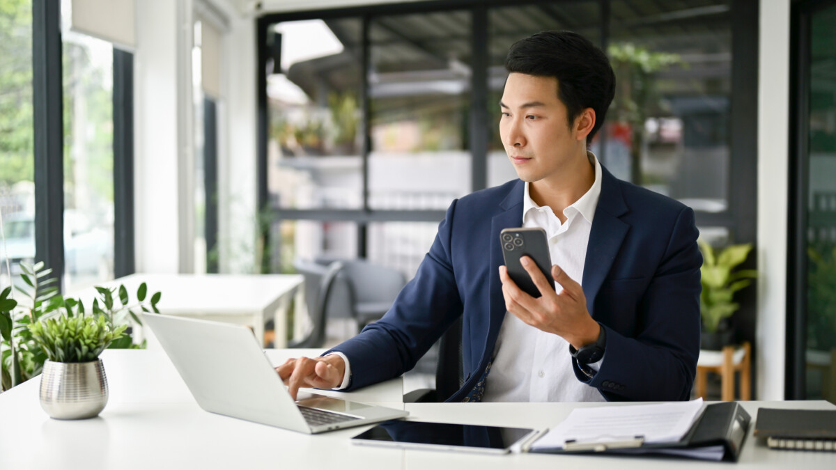 businessman holding his smartphone and using laptop at his desk