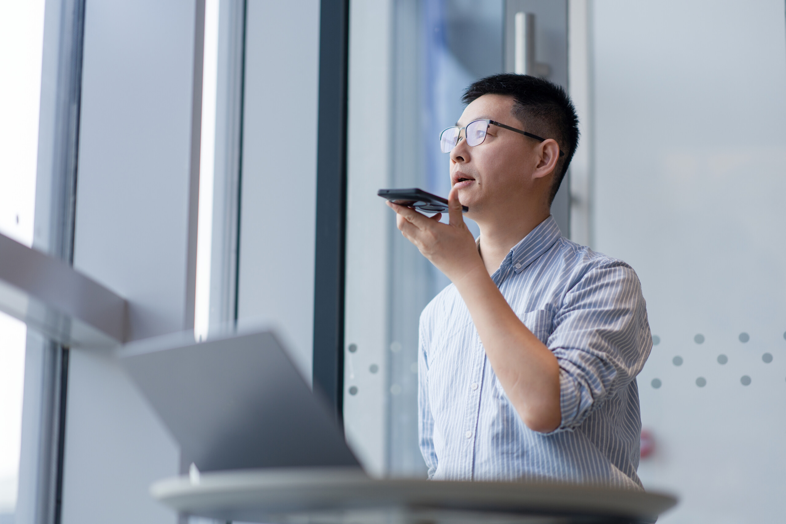 A man holds a smartphone and speaks voice commands while working in an office setting
