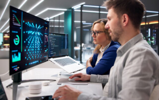 Two IT professionals analyze data on multiple screens in a modern server-room office.
