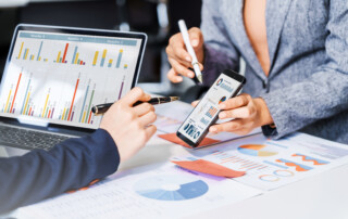Two female business professionals in suits sit at desk, engaged in strategic discussion. They analyze financial planning, risk management, corporate growth while reviewing reports and market trends.